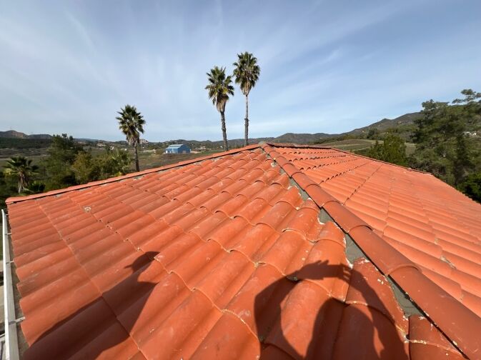 Red clay tile roof with visible ridge and shadows in the foreground; palm trees and distant hills in the background under a clear sky.