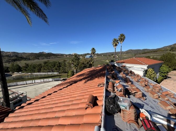 A red tiled roof under repair with stacks of tiles, tools, and materials, set against a backdrop of palm trees, hills, and a clear blue sky.