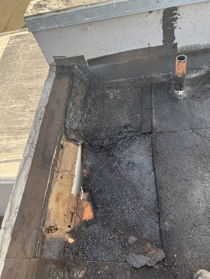 Damaged flat roof corner showing exposed insulation, deteriorated roofing material, and a small pipe protruding from the surface.