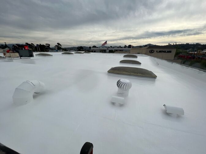 Flat white rooftop with multiple vents, skylights, and structures under a cloudy sky, with buildings and an American flag visible in the background.