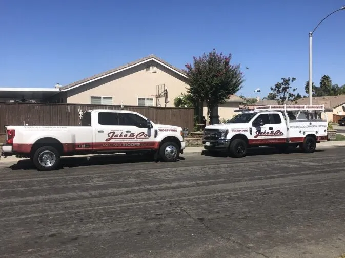 Two white pickup trucks with Jake & Co. branding are parked on a residential street in front of a house with a brown fence