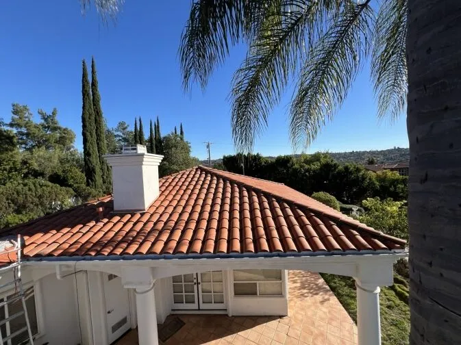 A single-story house with a red tile roof, white chimney, and white columns, surrounded by trees and under a clear blue sky.