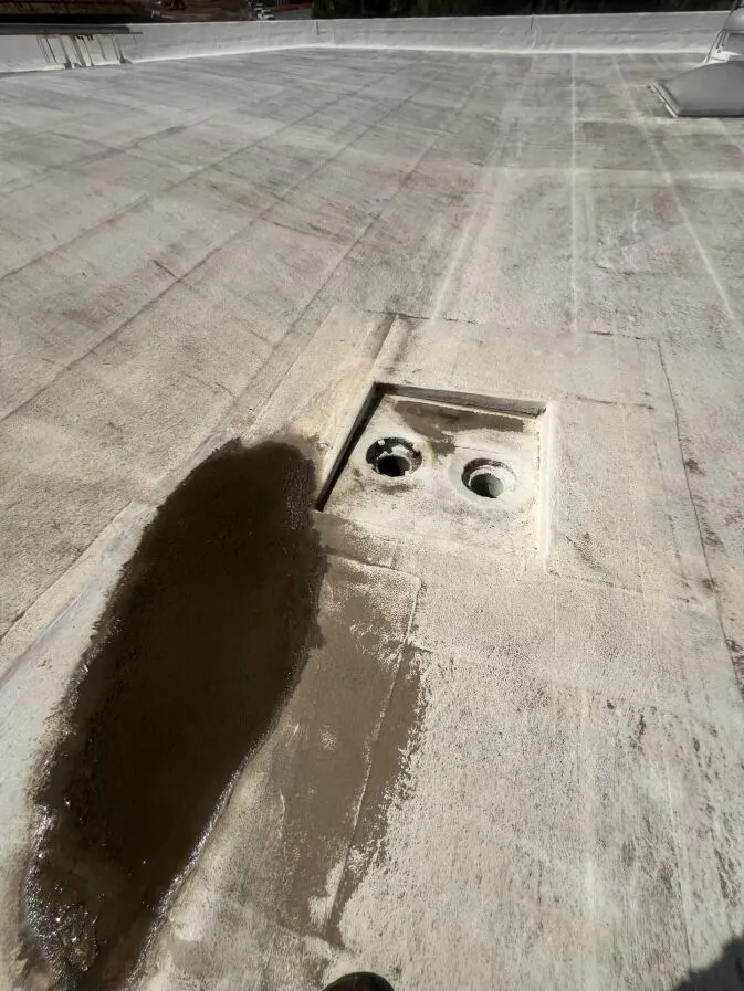 A flat rooftop with a water puddle next to an open drainage access panel containing two exposed drains.