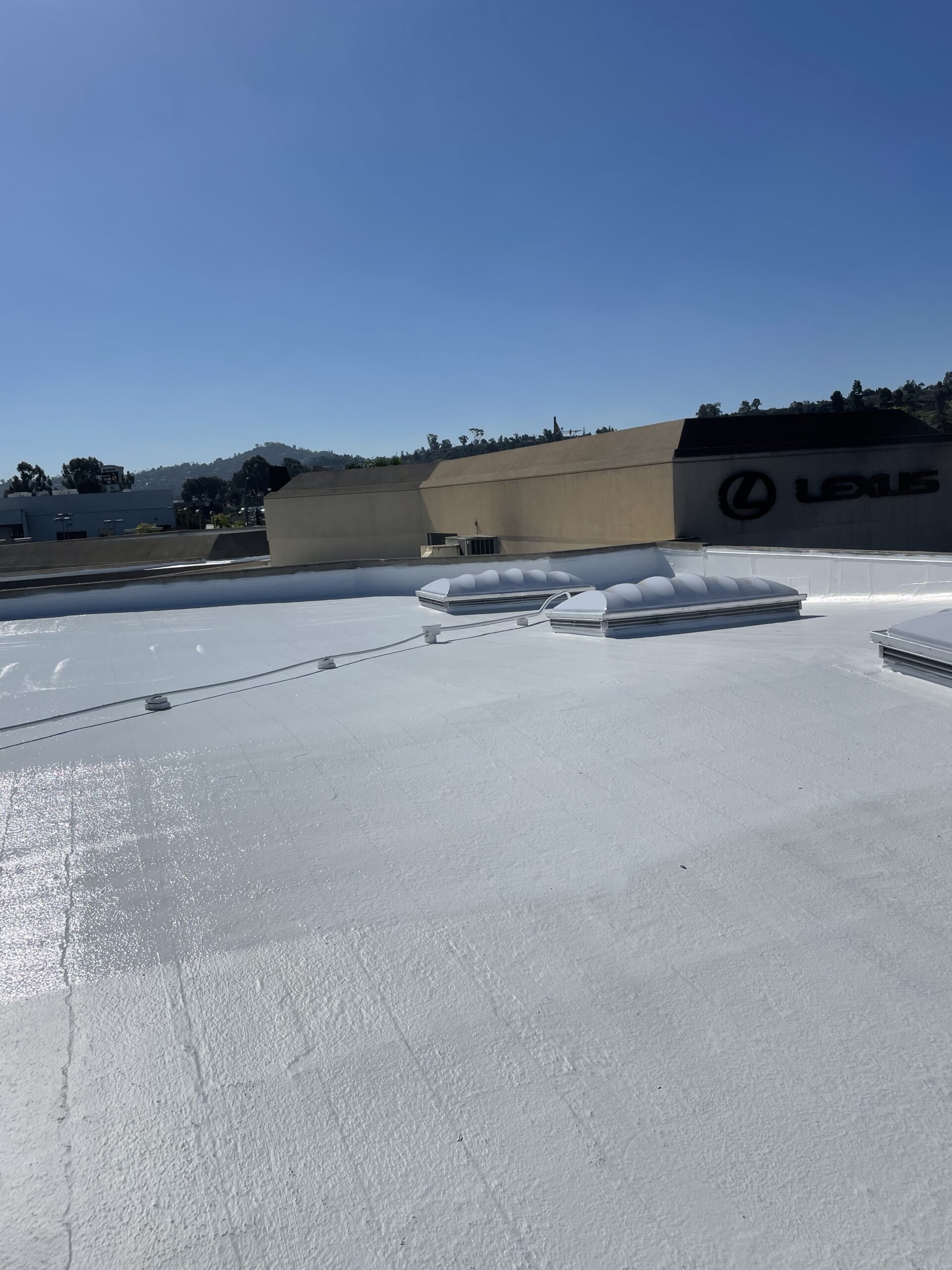 Rooftop with a white reflective coating and two skylights; Lexus dealership building and hills visible in the background under a clear blue sky.