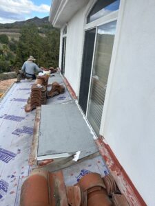 Worker installing roofing tiles on a flat section of a white building's roof, with stacked tiles and construction materials visible.