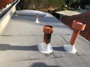 A flat rooftop with three orange ventilation pipes and a skylight, all bases sealed with white material; nearby are trees and red-tiled roofs.