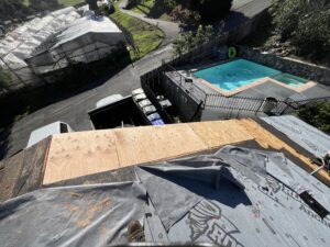 A rooftop under construction with plywood sheeting and roofing materials, overlooking a backyard with a pool and a road next to greenhouses.