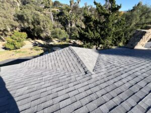 A roof with gray asphalt shingles shown from above, overlooking a yard with trees and greenery in the background on a sunny day.