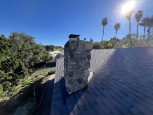 A stone chimney rises from a shingled roof under a clear blue sky, with tall palm trees and sunlight in the background.