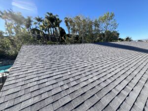 Gray asphalt shingles cover a sloped roof under a clear blue sky, with trees and a swimming pool visible in the background.