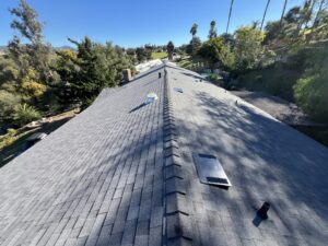 View of a gray shingle roof with several vents in a residential area surrounded by trees and greenery under a clear blue sky.