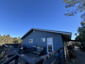A blue wooden house with a deck, outdoor furniture, and barbecue grill under a clear blue sky, surrounded by trees and plants.