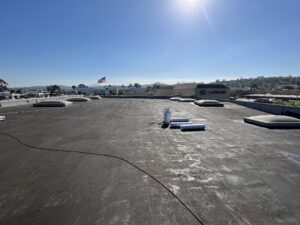 Flat commercial rooftop with skylights, HVAC equipment, and rolled roofing materials; clear sky and sun overhead; American flag and Lexus dealership in the background.