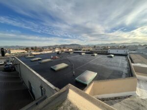 Flat commercial rooftop with multiple rectangular skylights, a few pieces of equipment, and a scenic view of distant hills under a partly cloudy sky.