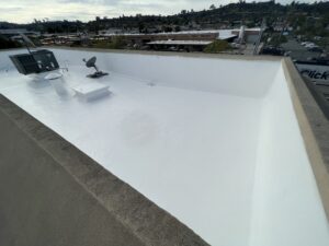 A flat commercial rooftop with a white reflective coating, HVAC unit, skylight, and satellite dish, surrounded by industrial buildings and a hilly landscape in the background.