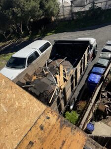 A truck with wooden side panels is filled with demolished roofing materials, parked beside a white vehicle and trash bins in a residential driveway.