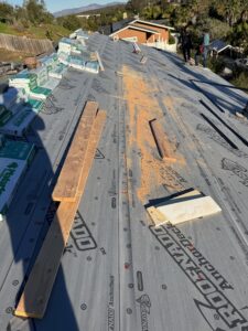 Stacks of roofing materials, wooden boards, and sawdust are scattered across a house roof under construction, with a few people standing in the background.