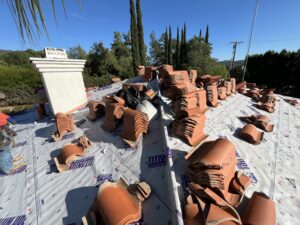 Stacks of clay roof tiles are arranged on a roof covered with synthetic underlayment, surrounded by trees and clear blue sky in the background.