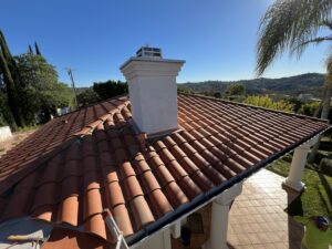 Red tiled roof with a white chimney in the center, viewed from above; palm tree and hills visible in the background under a clear blue sky.