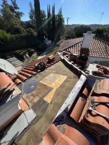 A rooftop under repair with exposed plywood and roofing material, surrounded by stacks of red tiles and tools on a sunny day.