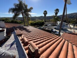A person works on a red tile roof under a clear sky, with palm trees, hills, vehicles, and houses visible in the background.