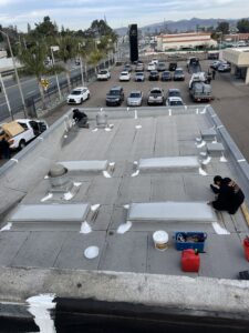 Workers apply sealant to vents and edges on a commercial building's flat rooftop, with a car dealership and parked vehicles visible in the background.