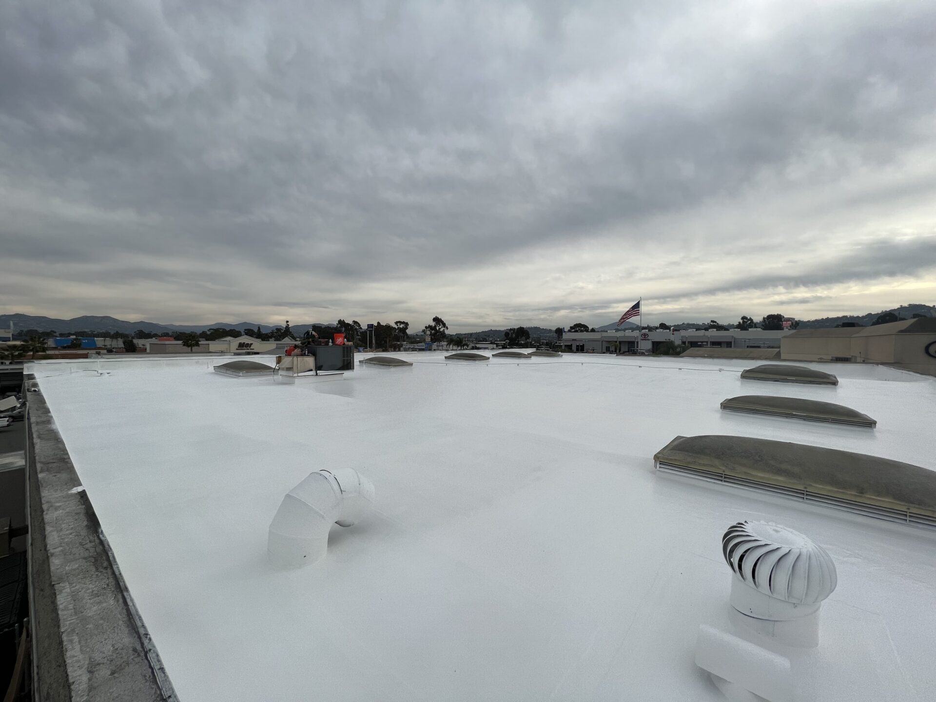 A flat white rooftop with several vents and skylights under a cloudy sky, with buildings and distant hills visible on the horizon.