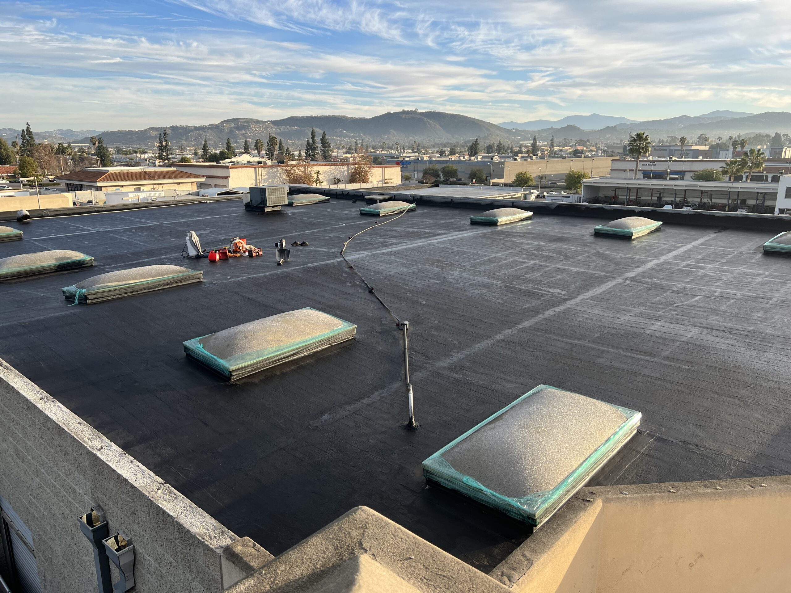 Flat rooftop with several covered skylights, construction materials, and two workers; surrounding buildings and mountains visible in the background under a blue sky.