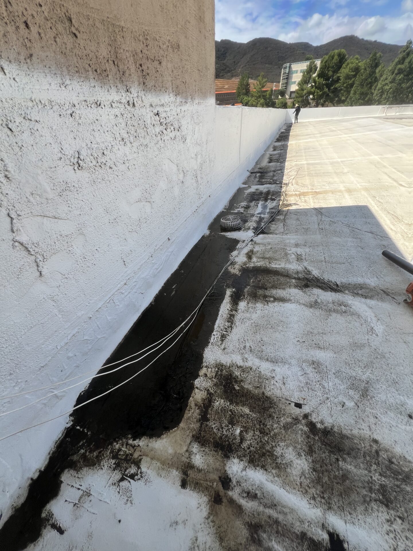 A rooftop with a white painted wall, visible drainage grate, and dark water stains along the base. Hills and buildings are seen in the background.
