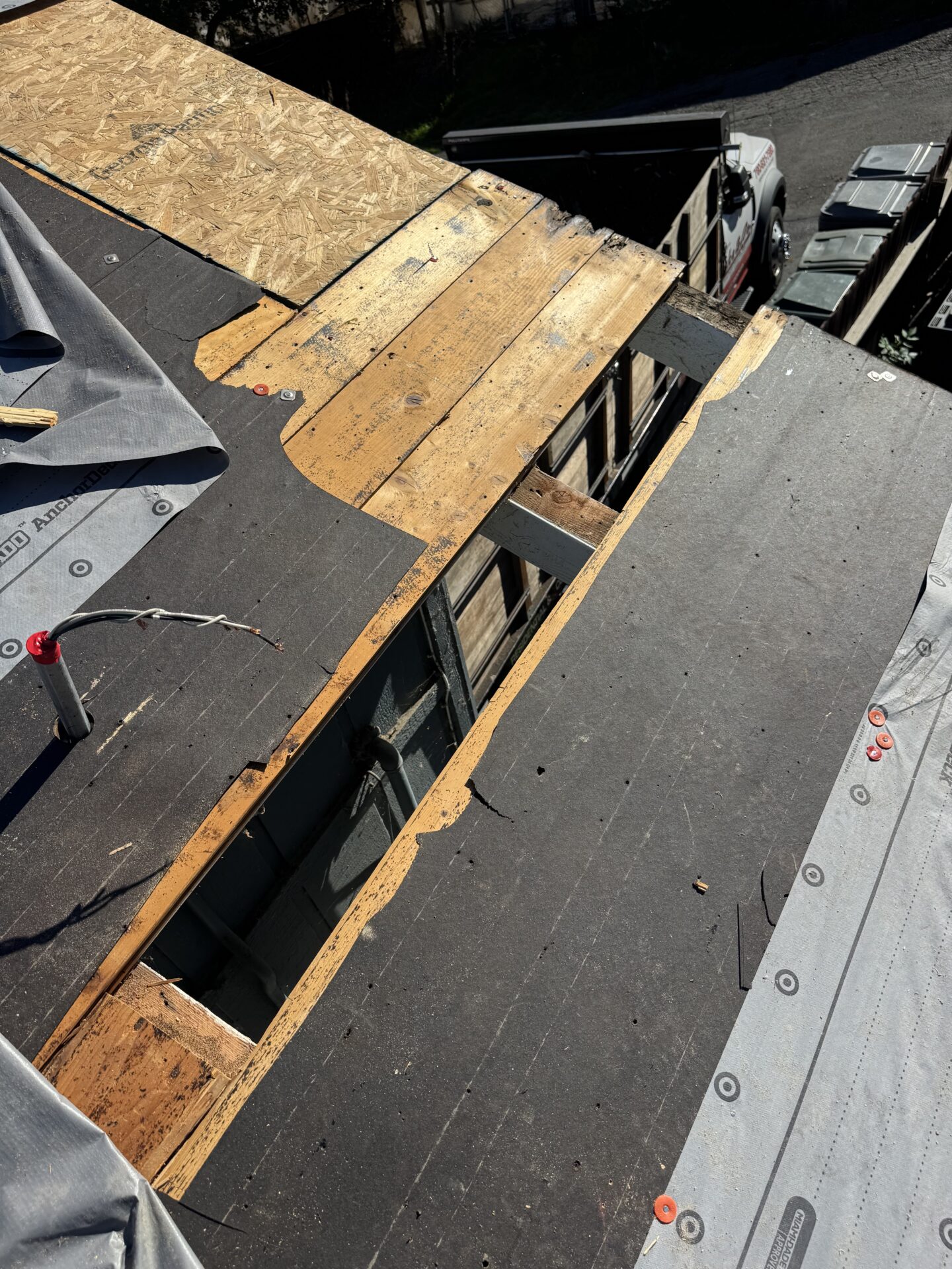 Partially removed roof sheathing exposing wooden beams and underlayment during roof repair or replacement; tools and materials are visible on the roof.