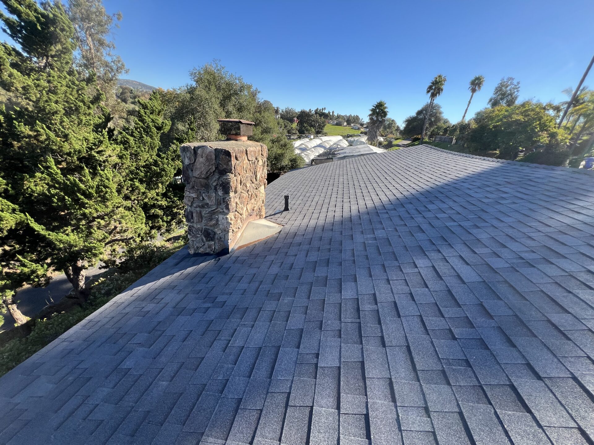 A shingled roof with a stone chimney, surrounded by trees and greenery under a clear blue sky.